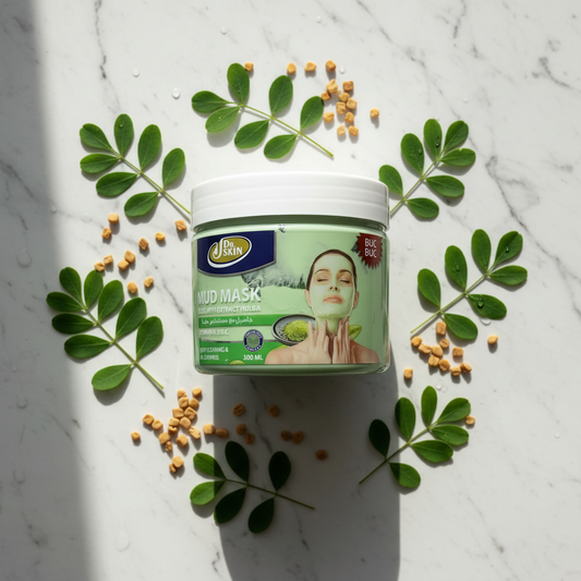 A tub of Johnnie Green Mud Mask centered on a white marble surface, surrounded by green leaves and small tan seed pieces in a styled flat-lay arrangement.