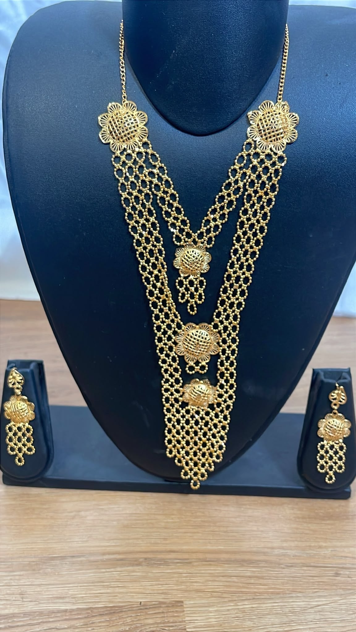 Gold-toned traditional Indian jewelry set featuring a multi-strand chain necklace with ornate floral and bead motifs, displayed on a black mannequin bust with matching drop earrings on small stands against a light wood surface.