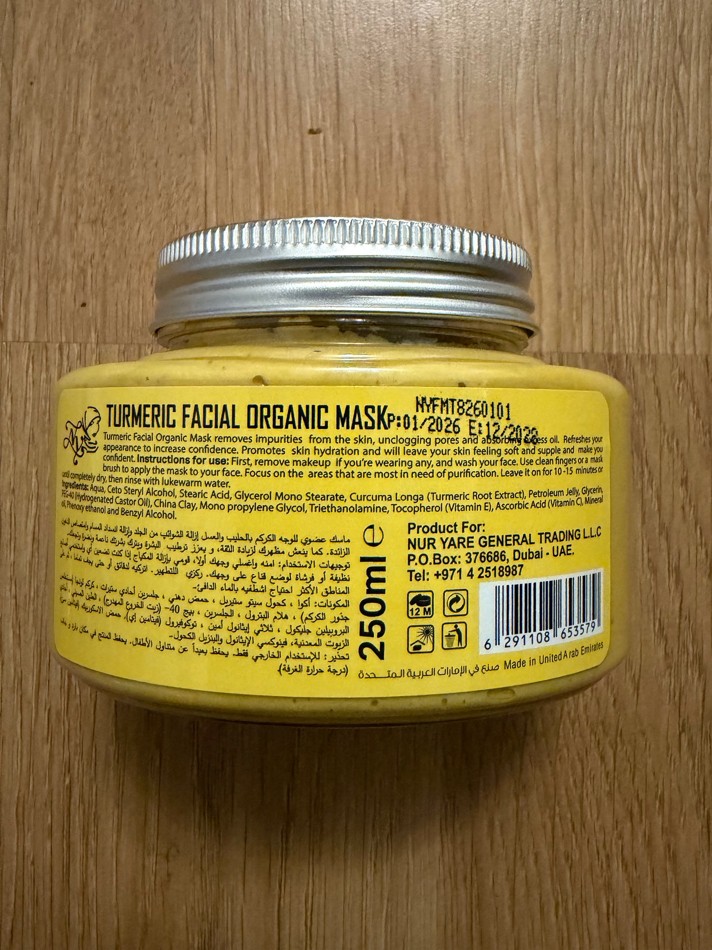 A yellow jar of Turmeric Facial Organic Mask by Nu Ye General Trading LLC, showing the back of the label with product details, a silver screw cap, and a barcode, placed on a wooden surface.