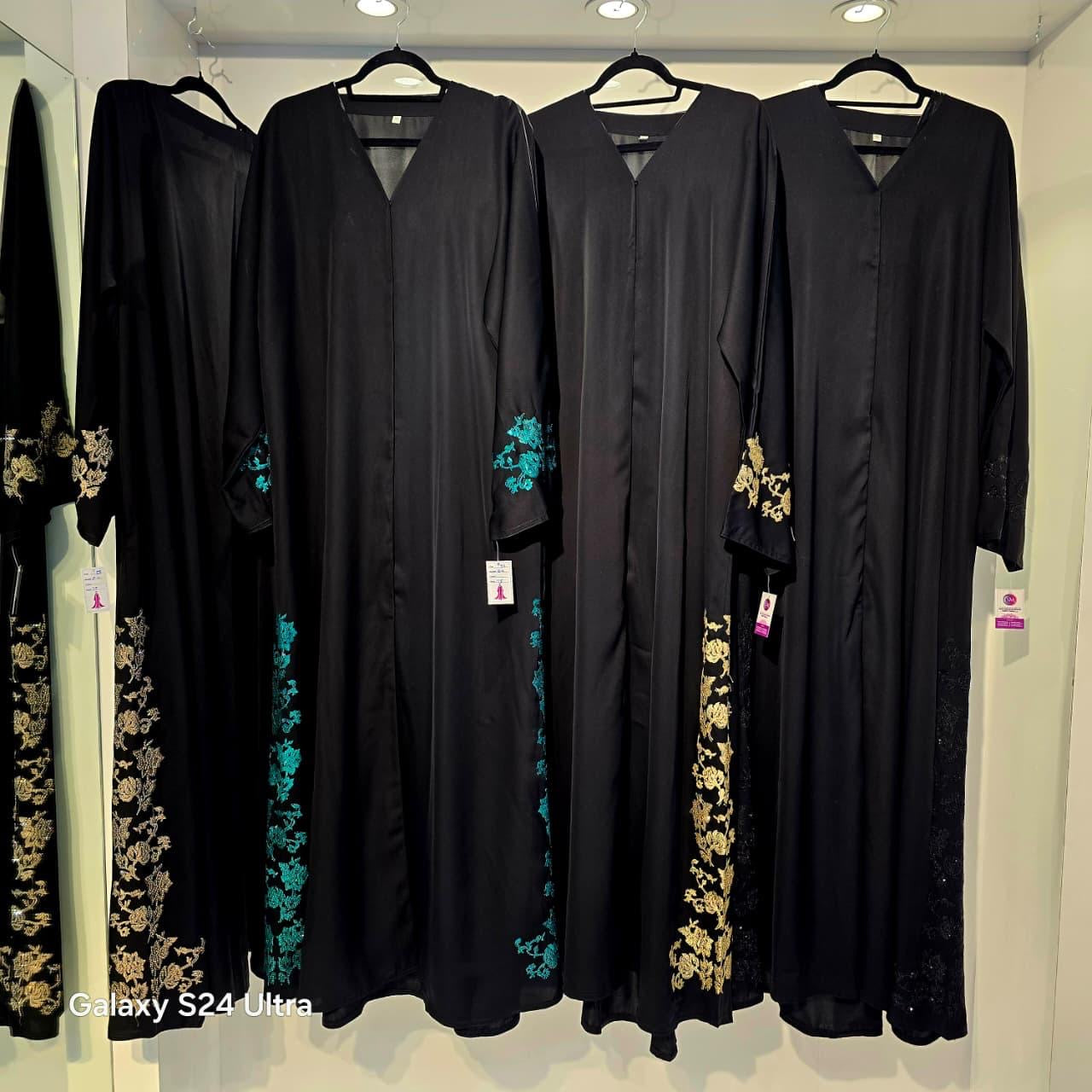 Black abayas with floral embroidery on hangers against a white wall.