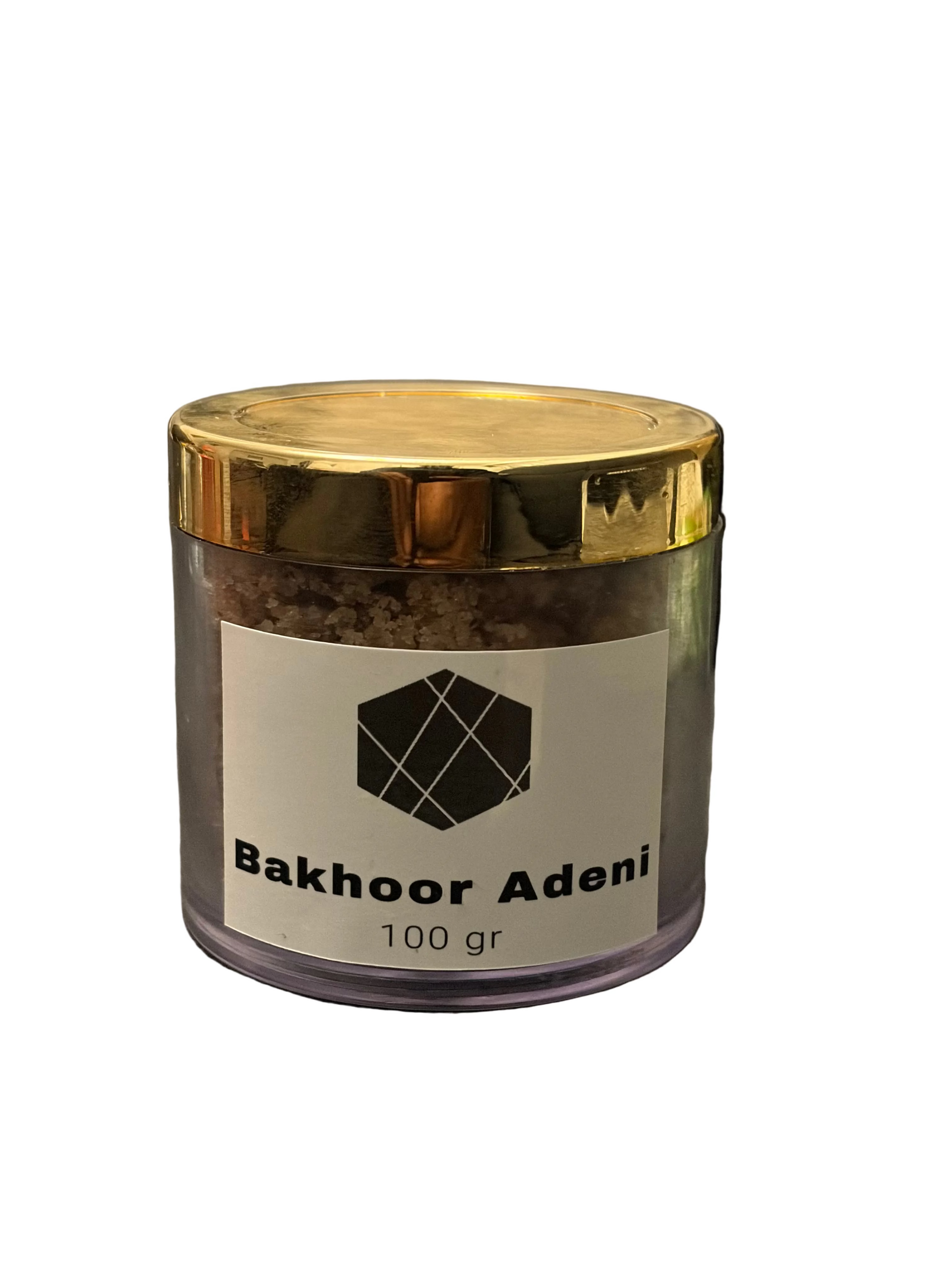 A clear plastic jar of Bakhoor Adeni incense with a shiny gold lid and a white label, placed on a dark gray textured surface with a blurred laptop in the background.