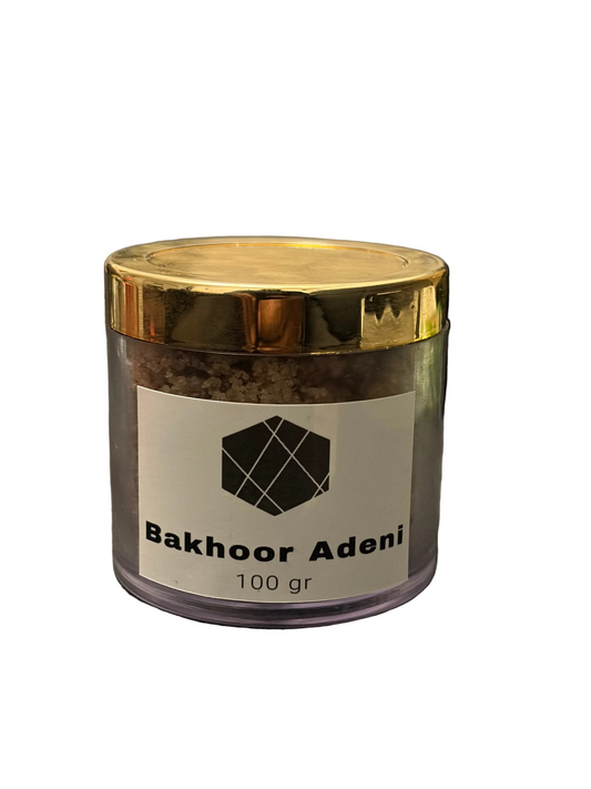 A clear plastic jar of Bakhoor Adeni incense with a shiny gold lid and a white label, placed on a dark gray textured surface with a blurred laptop in the background.