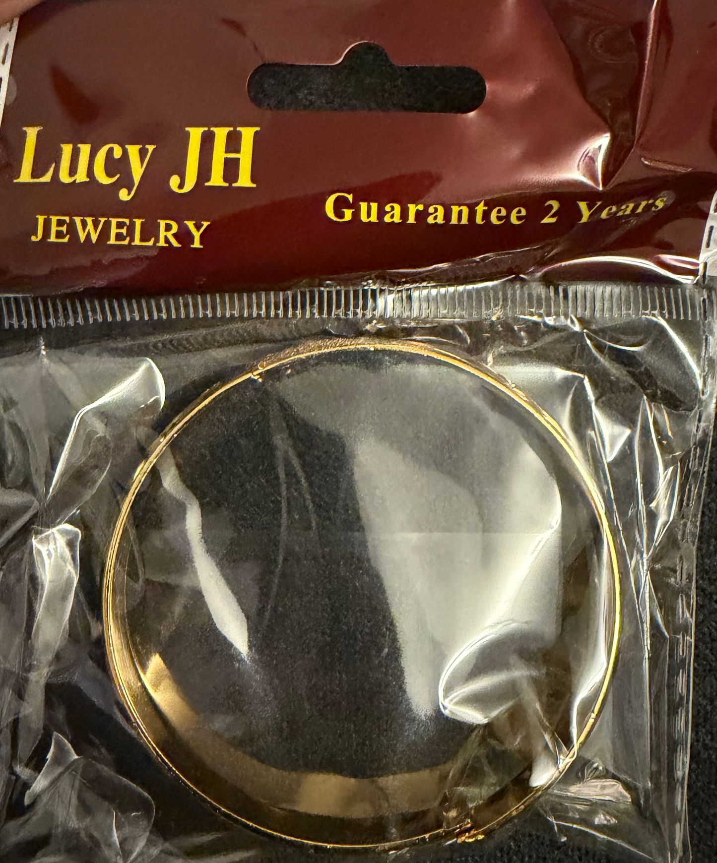 Gold circular object in clear packaging with 'Lucy JH Jewelry' label.