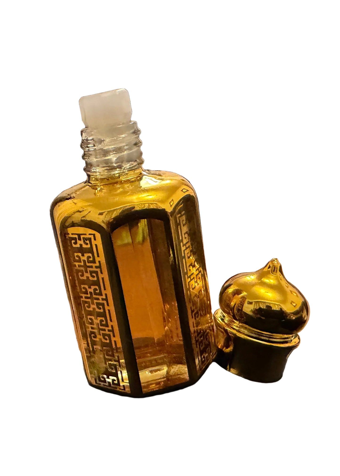 A small rectangular glass bottle filled with golden-yellow liquid, shown with its matching gold cap beside it on a black surface. The background is a cluttered indoor room with a chair, door, and boxes.