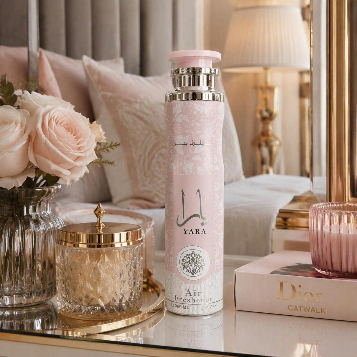A pink and white Yara Air Freshener bottle with silver accents, styled on a glass table next to a Dior book, a pink candle, and a vase of roses in a luxurious bedroom setting.