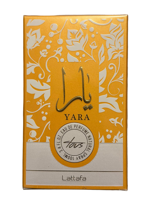 A rectangular yellow and white Lattafa Yara perfume box placed on a wooden surface. The box features white floral patterns, Arabic calligraphy, and a circular logo for Tous.