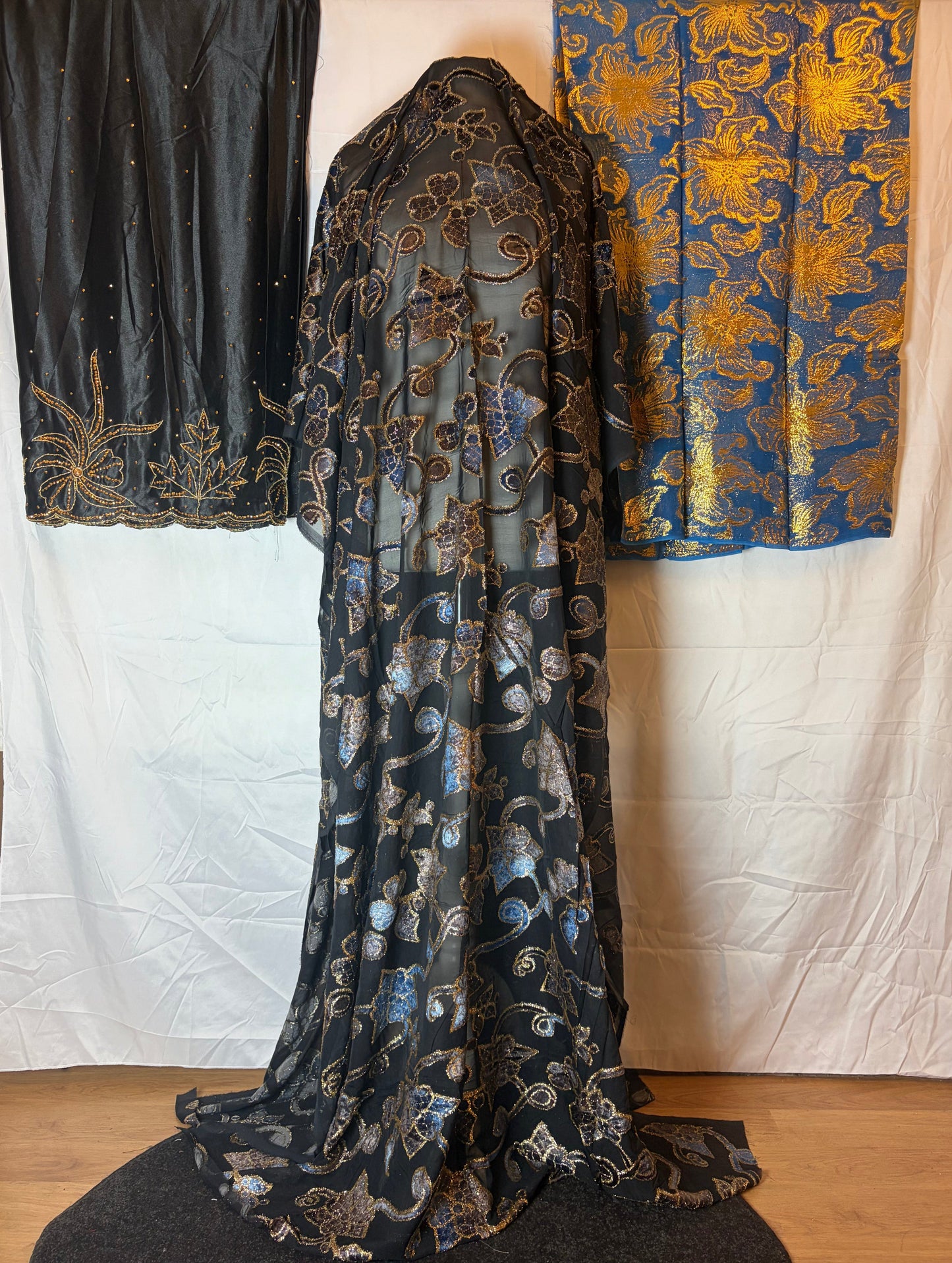 Decorative black fabric with gold patterns draped over a white surface, with two other fabrics in the background.
