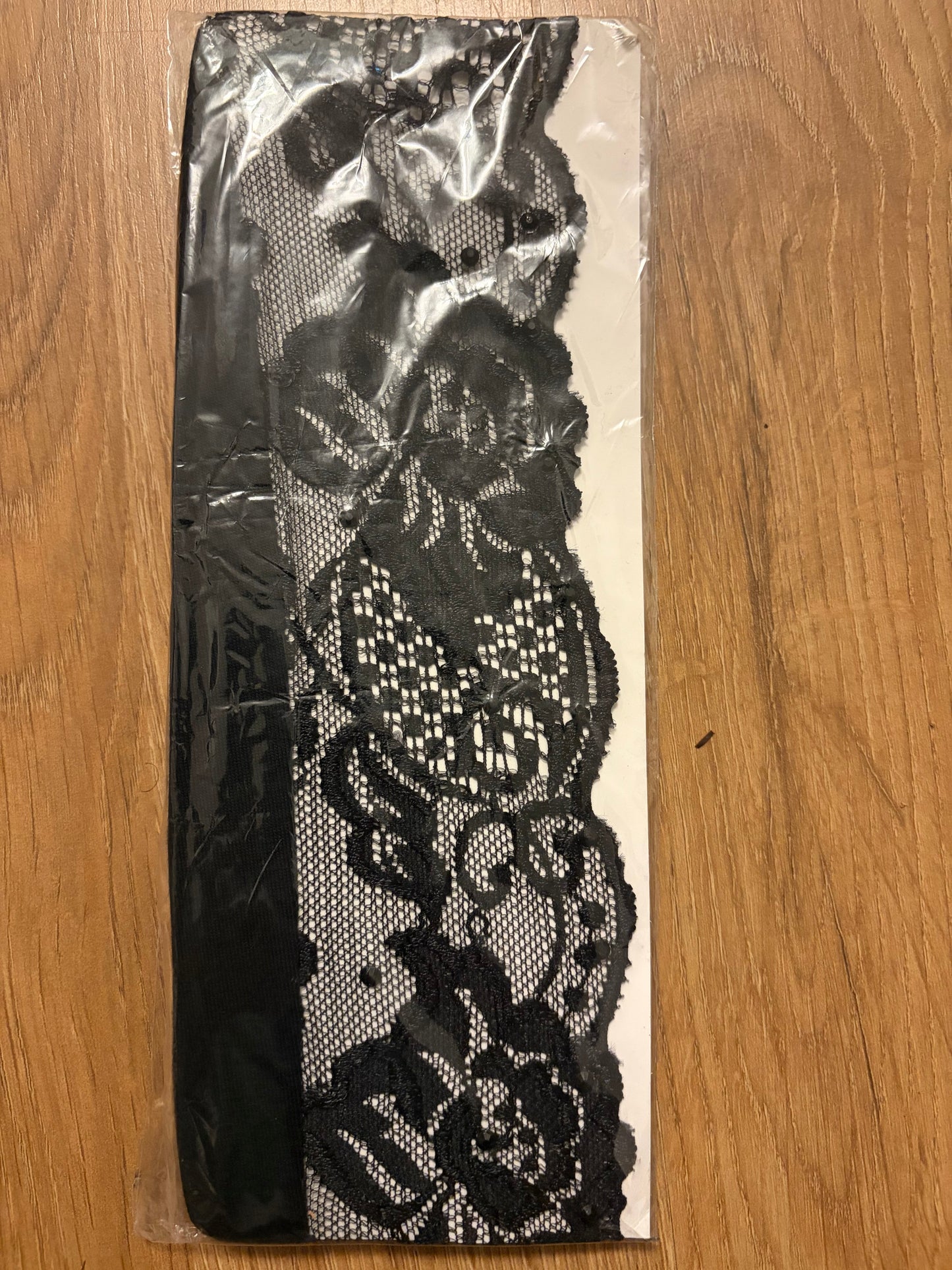 Black floral lace trim with a sheer mesh center, packaged on a white backing sheet and placed on a wooden surface.