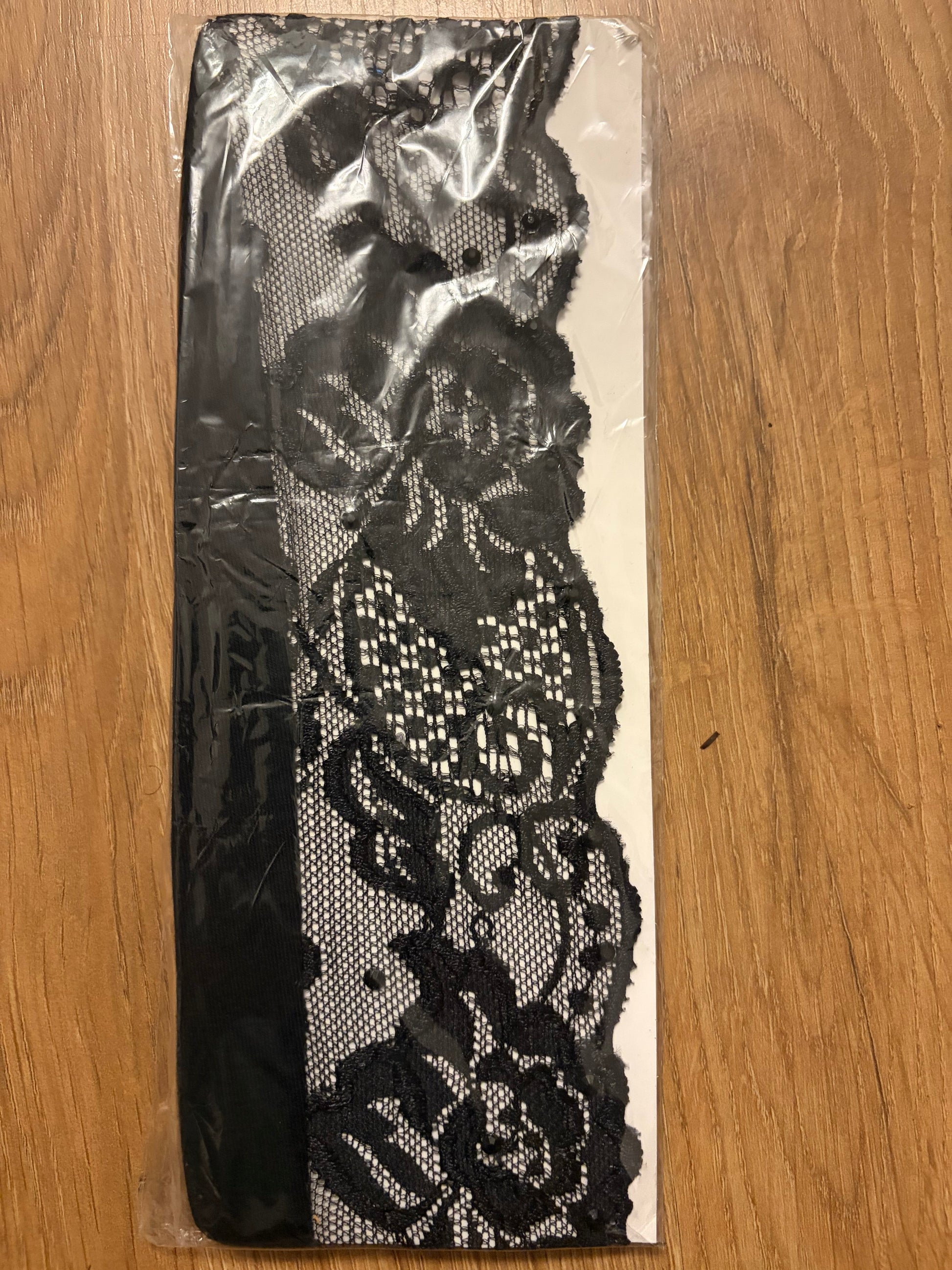 Black floral lace trim with a sheer mesh backing, wrapped in clear plastic packaging and placed on a wooden surface.