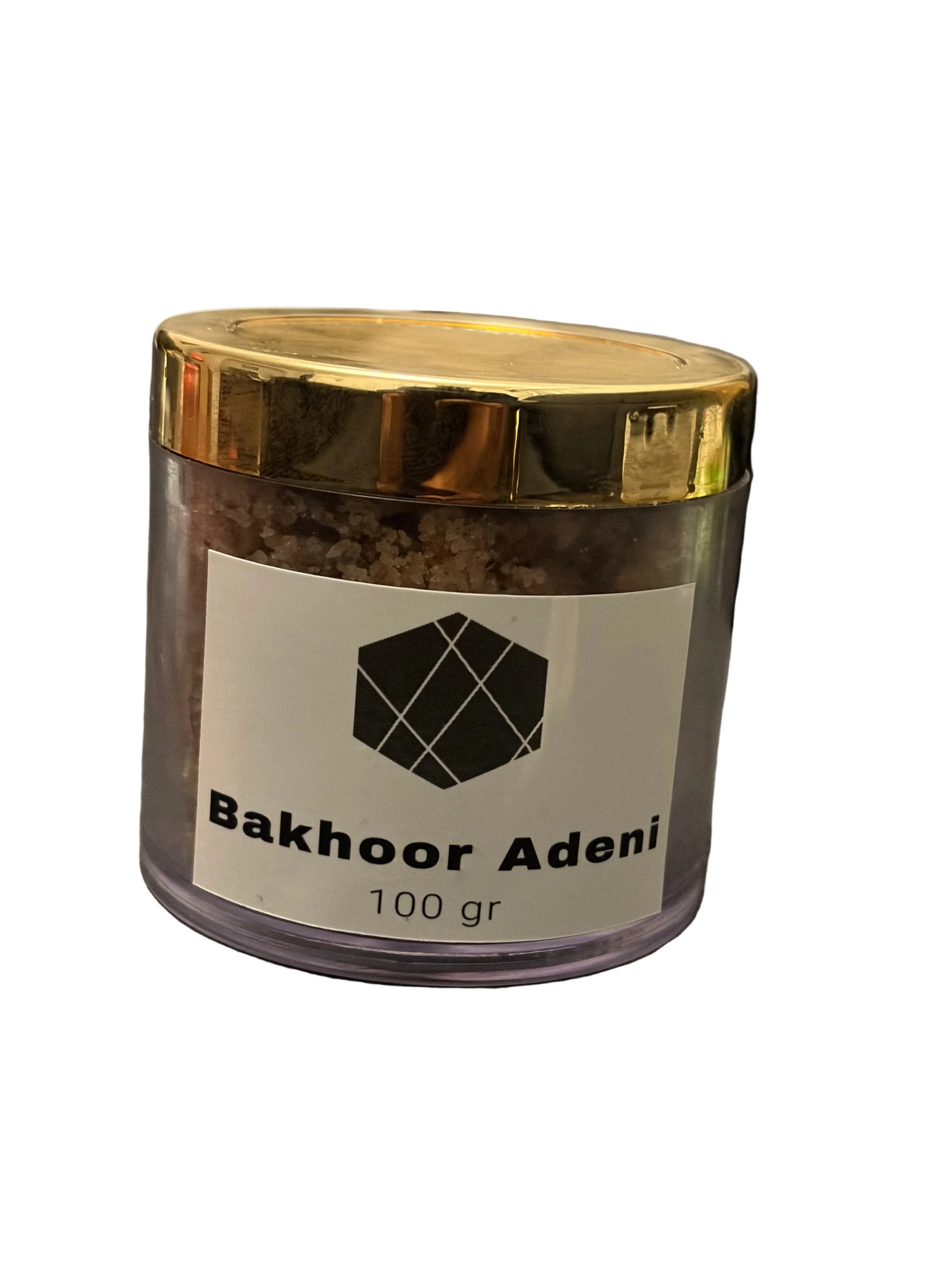 A clear plastic jar of Bakhoor Adeni incense powder with a shiny gold lid and a white label, resting on a dark gray fabric surface.
